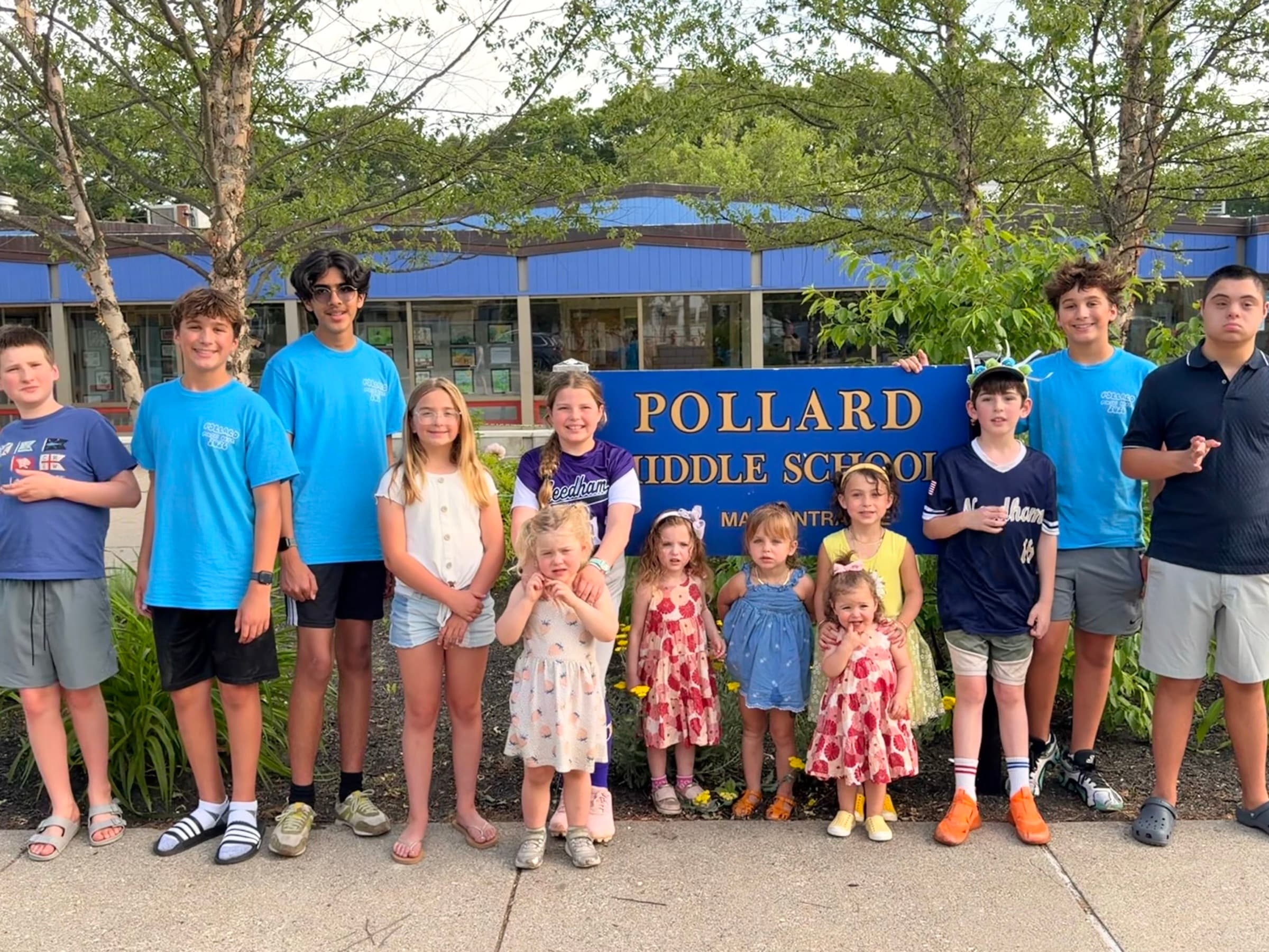About a dozen Needham kids of all ages gathered together in front of the Pollard Middle School entrance sign on a summer evening.