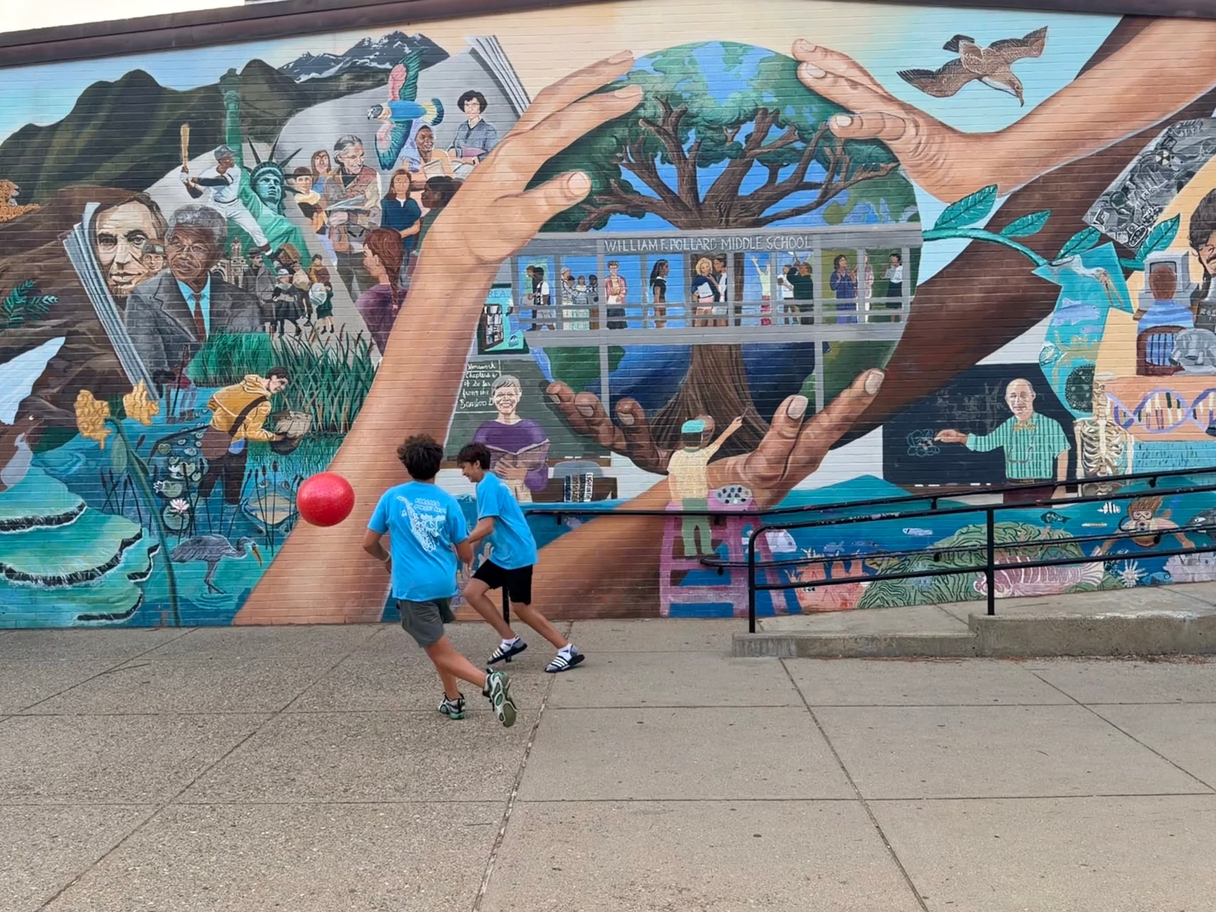 Two kids playing with a red ball in front of Pollard’s colorful community mural of hands cradling a tree.
