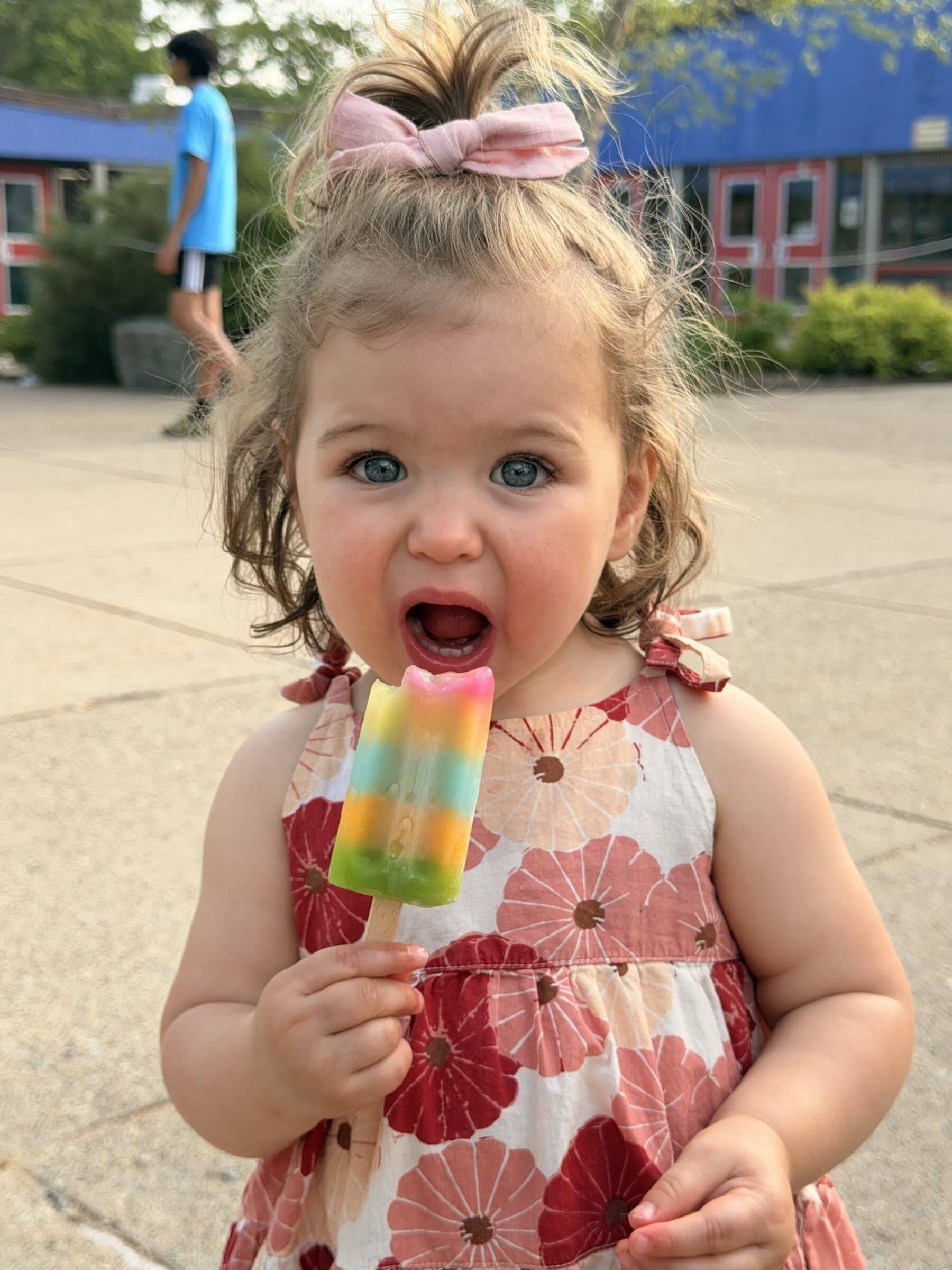 A young child in a floral dress happily eating a rainbow popsicle outside Pollard Middle School.