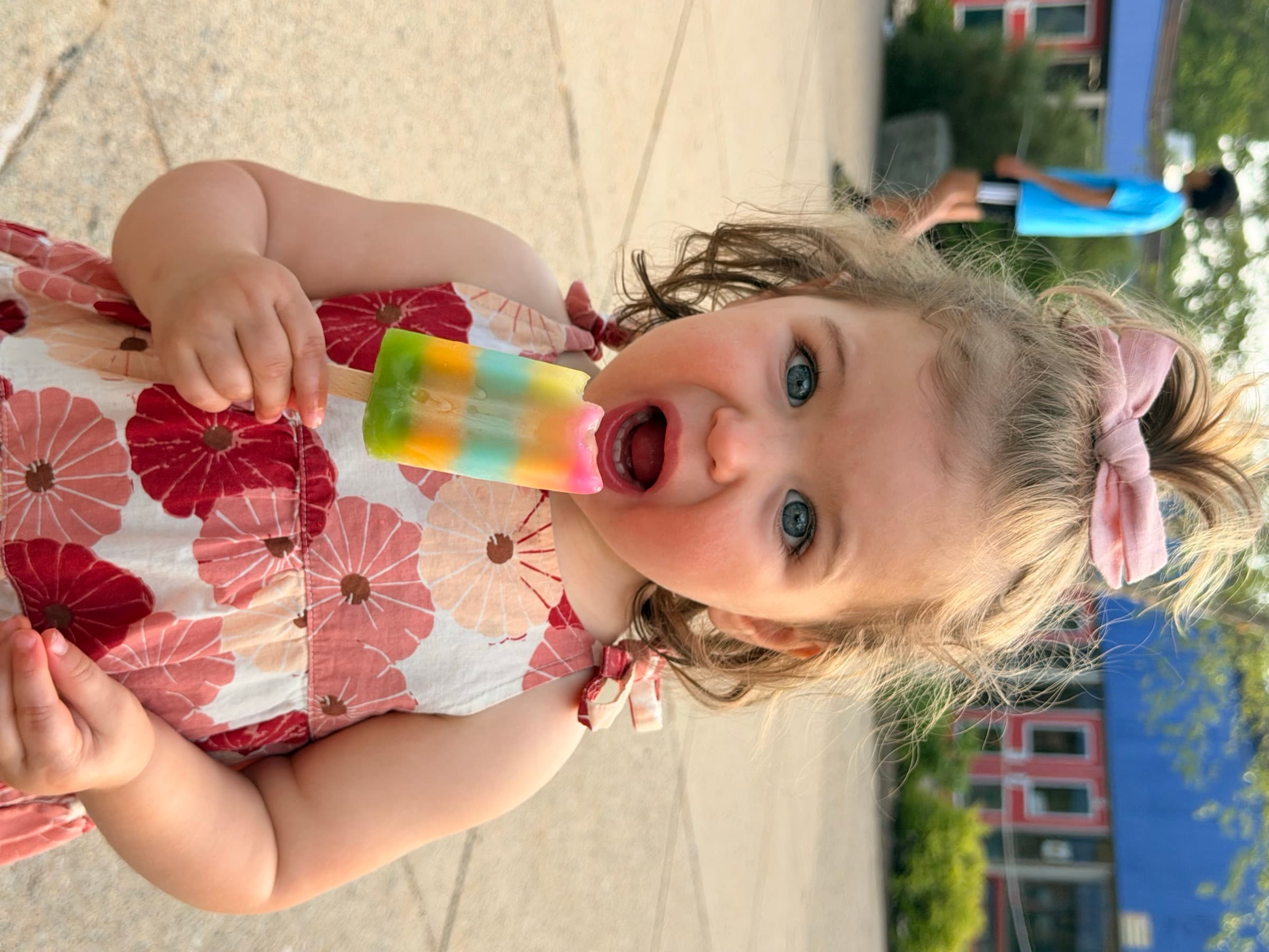 A young child in a floral dress happily eating a rainbow popsicle outside Pollard Middle School.